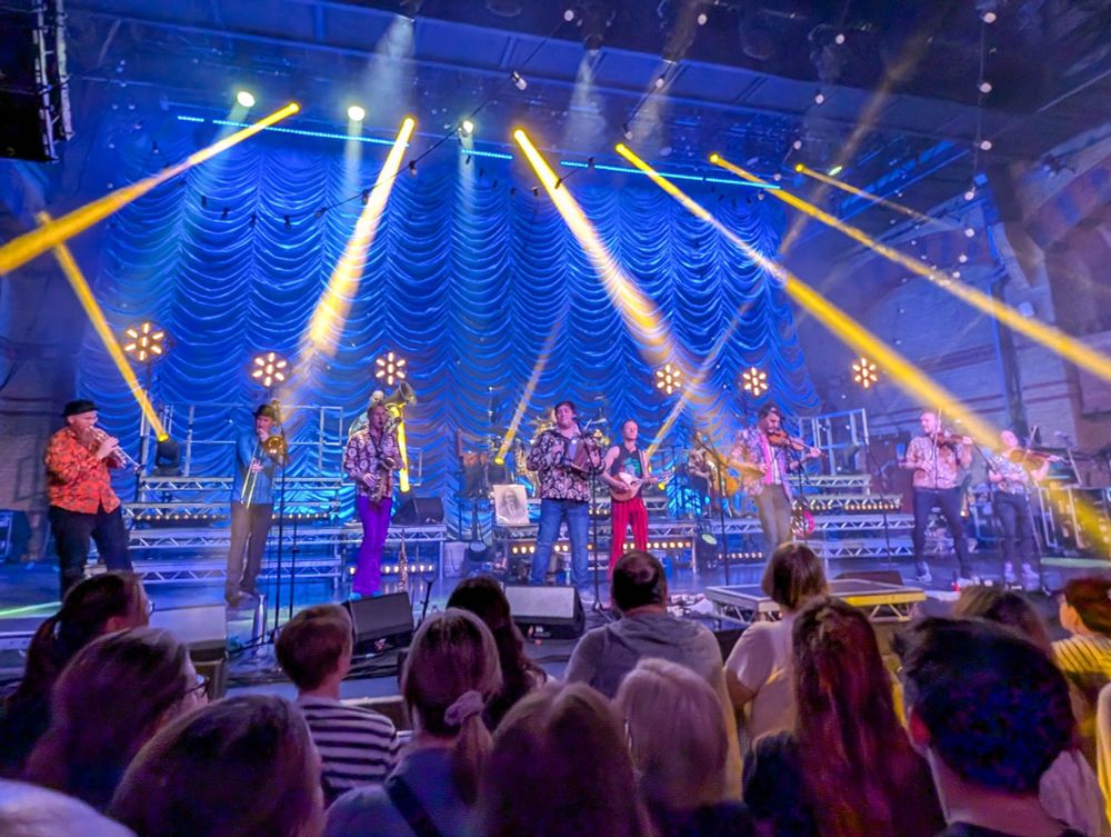 Bellowhead on stage with flowery clothes and beams of light.