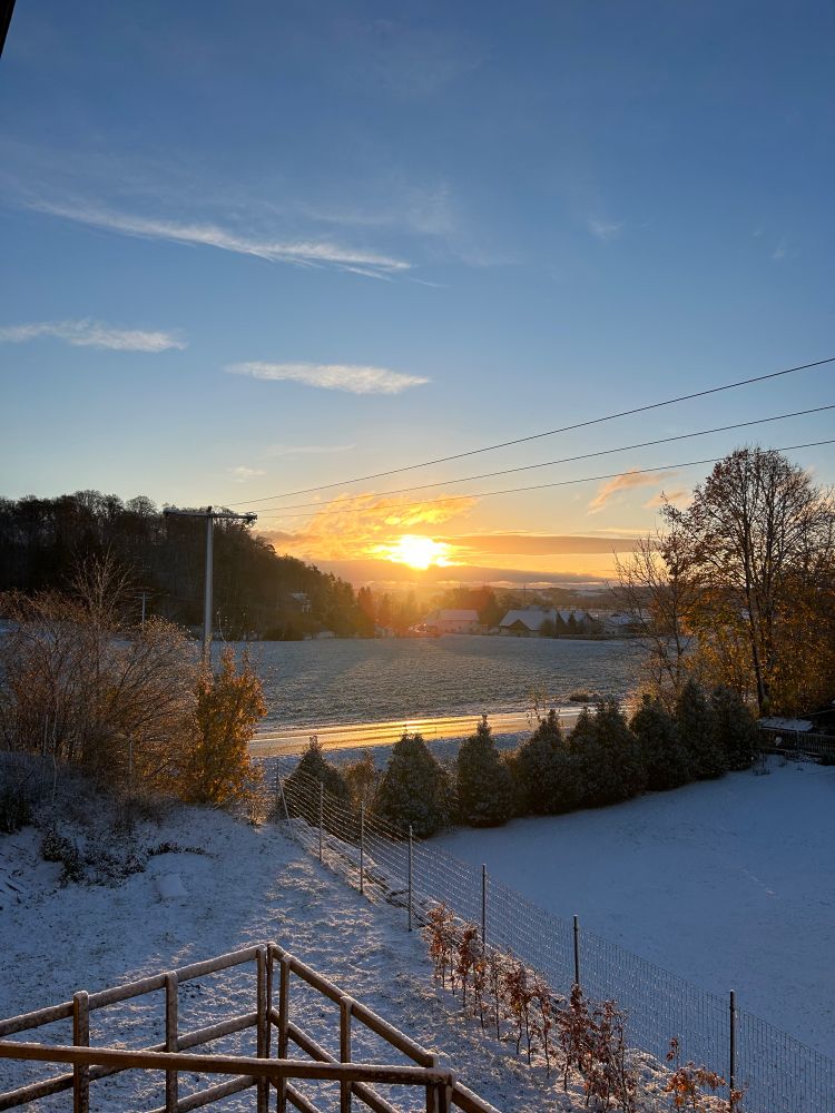 Schnee liegt auf dem Rasen und den Feldern während im Hintergrund die Sonne langsam aufgeht.