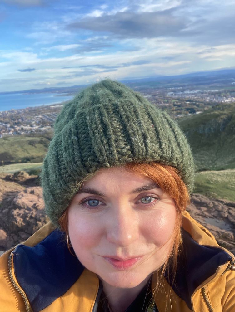 A selfie of me, a white 36 y/o woman. I’m wearing a green, knitted beanie, a yellow coat with blue collar and you can see a few strands of red hair. Behind me is the view from Arthur’s Sea, it’s mostly free, you can see in the distance Edinburgh and a bit of the sea