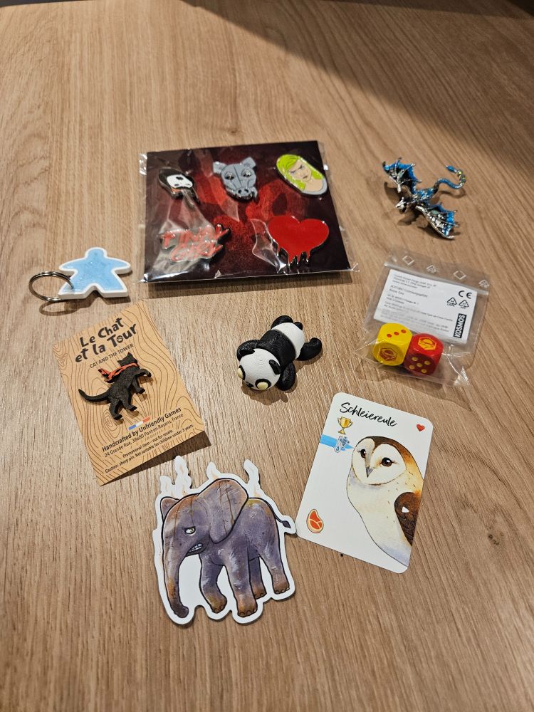 Kostenlose Beigaben der SPIEL:
- Final Girl Pins
- ein Meepleschlüsselanhänger
- ein Metalldrachen-Pin
- ein Holzkatzenpin
- ein 3D-gedruckter Panda
- 2 Würfel 
- eine Schleiereulen-Karte
- ein Elefantenmagnet