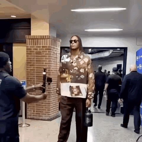 a man in a floral shirt is standing in a hallway holding a water bottle while being interviewed .