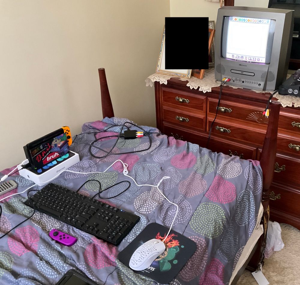 The setup I used for the Mario Paint attack; a mouse, keyboard and Joycon (to navigate other menus) are there for me to use, the Switch in docked mode is on the bed standing on a box to make sure it balances (and to ensure I have a mouse to use this with), and a HDMI-to-AV converter on the bed that connects to the switch dock to the CRT tv.
