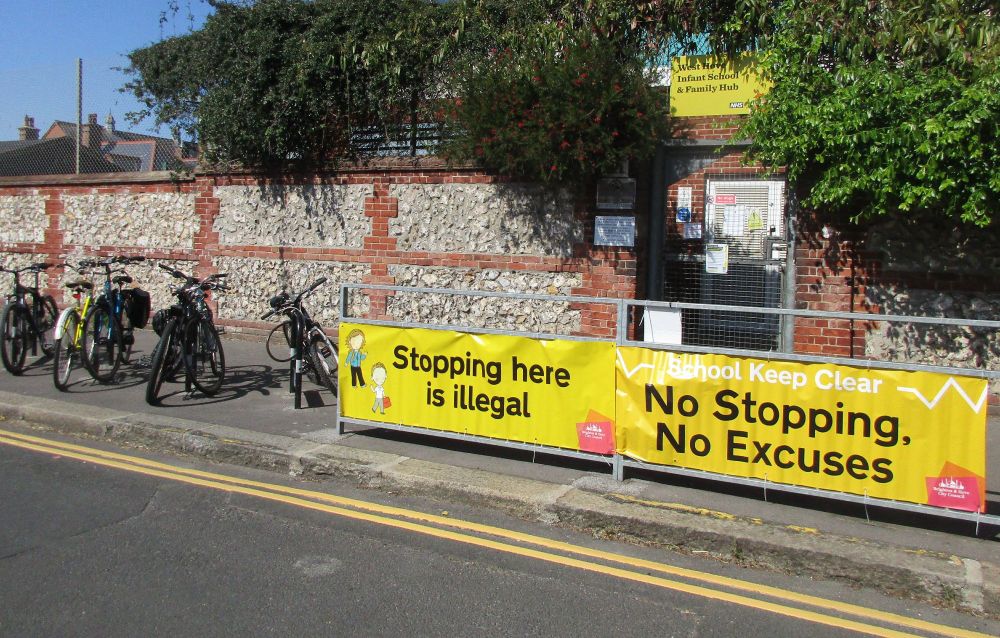 Banner outside a school street: "Stopping here is illegal. No Stopping. No Excuses." Bike parking on the left. Flint wall and gate behind.