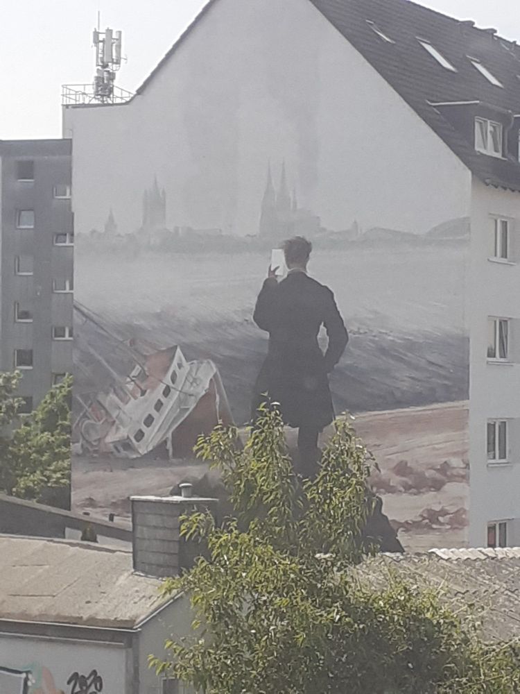 Auf einer Hauswand gemalt, angelehnt an Caspar David Friedrichs "Wanderer über dem Nebelmeer": eine mit dunklem Jackett gekleidete Figur, die Betrachtenden den Rücken kehrt. Sie hält in der linken Hand ein Blatt und scheint zu lesen. Im Horizont die Silhouette der Stadt Köln mit Dom, Rauch steigt aus einem Schornstein, das hier düstere Grautöne, Links, zu den Füßen der Figur, ein gestrandetes weiß-rotes Schiff vor hohem Wellengang, graublaues Meer.