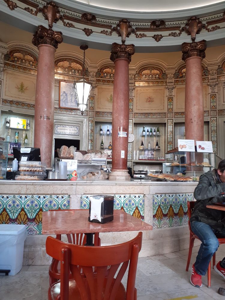 Tresen eines Cafés, heller Marmor mit Fliesen, die Blumen, Korn und Bienen abbilden; im Hintergrund drei rotrosa Marmorsäulen, wo sie an die Decke münden, bronzefarbene Kapitellverzierung; im Vordergrund ein rotrosa Marmortisch mit zwei Holzstühlen