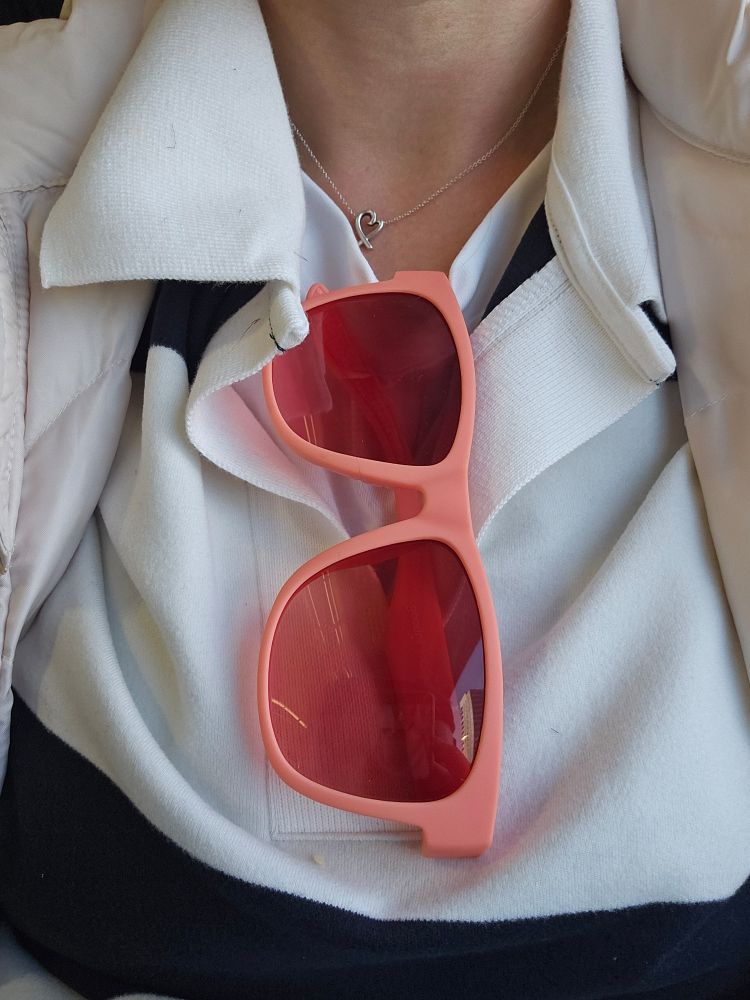 Pink framed sunglasses with pink lessons hanging from the neck od a blue and white top