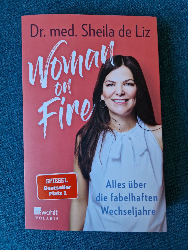 Ein rötliches Taschenbuch, auf dessen Titel das Foto einer hellhäutigen Frau mit langen, dunklen Haaren und einem weißen, ärmellosen Oberteil abgebildet ist. Der Titel lautet "Woman on Fire ‐ Alles über die fabelhaften Wechseljahre". Die Autorin ist Dr. med. Sheila de Liz, der Verlag ist Rowohlt Polaris. 