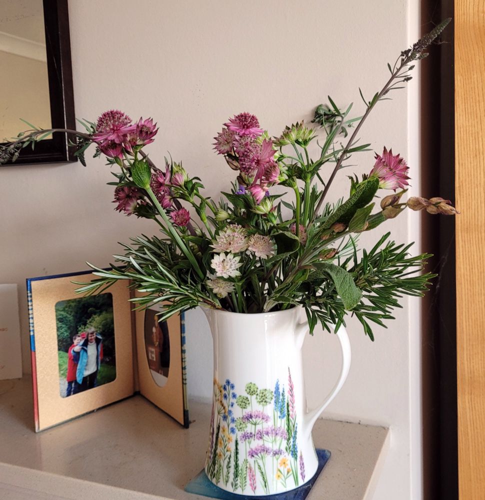Red, purple and white flowers, with greenery, in a jug-vase. On a mantelpiece.
