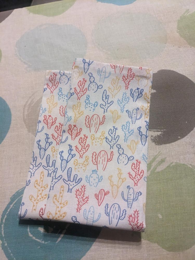 Two folded cloth handkerchiefs. They are white, with a colorful print motif of smiling cacti. The backdrop is an ironing board.
