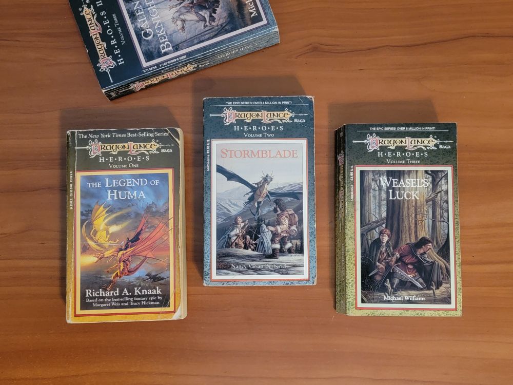 4 Dragonlance novels on a wooden desk: the complete Heroes trilogy, plus a book from the Heroes II trilogy, partially cut off in the image. The titles of the three Heroes I trilogy books are: The Legend of Huma, Stormblade, and Galen's Luck. There are dragons on the first two covers, and some adventurers hiding in the forest on the third. 