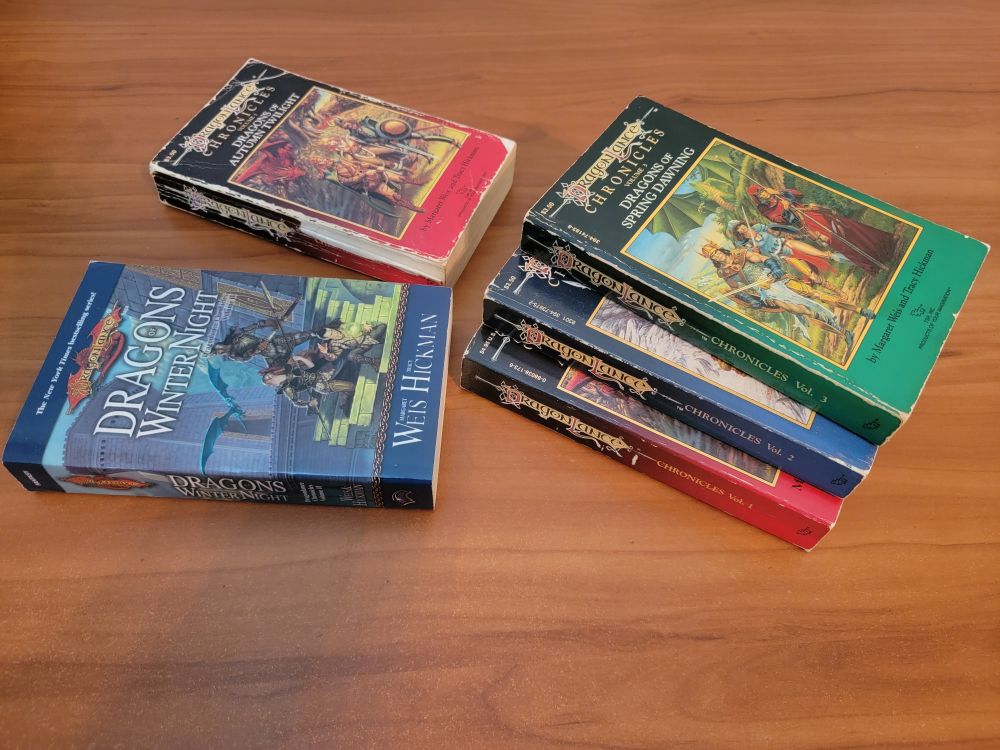 The Dragonlance Chronicles trilogy, first edition paperbacks, plus a tattered 1st ed Dragons of Autumn Twilight, and a pristine 90's second edition reprint of Dragons of Winter Night. A total of five fantasy novels in red, blue, and green, all with dragons on the front.