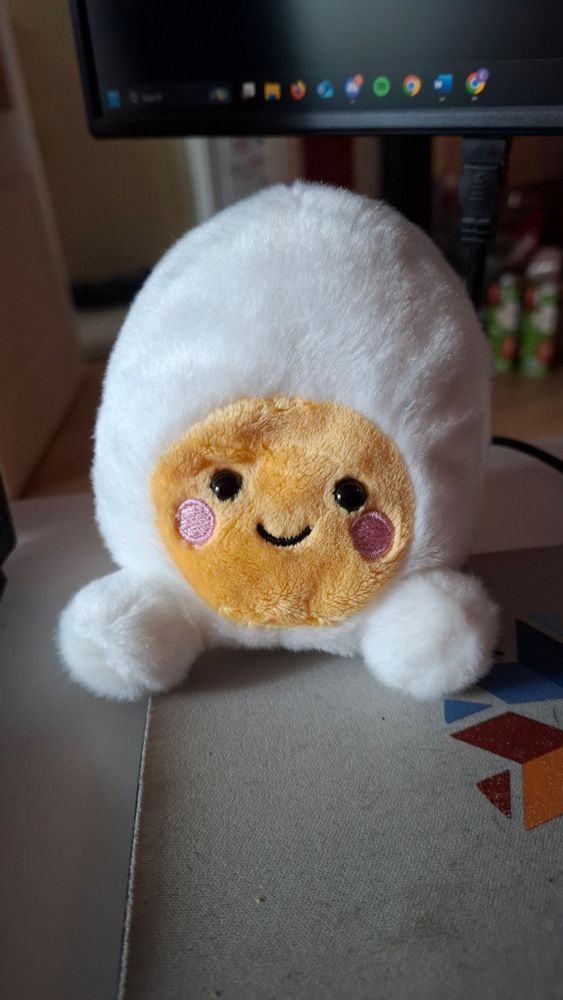 A small plushie of an egg with a smiley face. The whole body is a white egg with tiny round feet and no arms, and it has a flat yellow yolk face with a big happy smile and little pink rosy cheeks. It is sitting on my computer desk next to my mouse, and it is fucking adorable. 