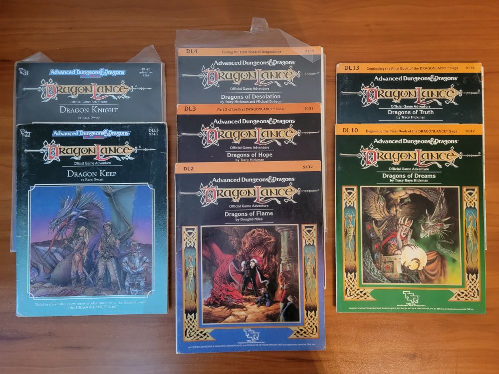 A collection of seven Dragonlance AD&D modules back from the old days, primarily by Tracy Hickman. DL2, DL3, DL4, DL10, DL13, and a couple other dragon-themed modules are shown on a wooden desk.