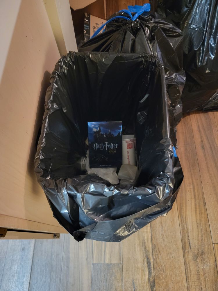 A Harry Potter DVD box set sits proudly in a kitchen trash can lined with a black trash bag, amidst other garbage, where it belongs.