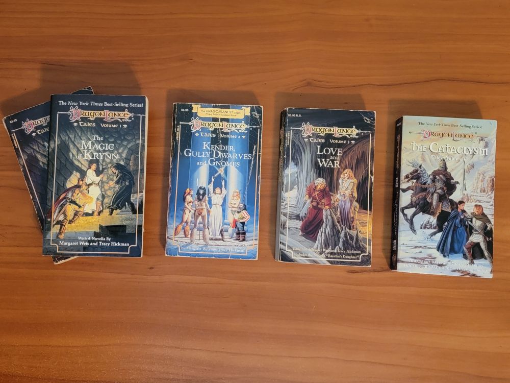 5 Dragonlance novels in a row on a wooden desk: the Tales I trilogy, plus the first book of Tales II. Book titles are The Magic of Krynn (x2), Kender Gully Dwarves and Gnomes, Love and War, and The Cataclysm. Books are mostly in good or very good condition. The duplicate of The Magic of Krynn is in rougher shape.