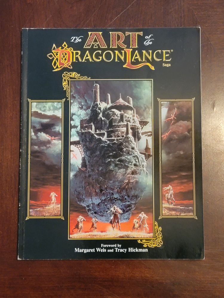 The book The Art of the Dragonlance Saga is on a glossy wood table. The book is the 90's second edition version with a black cover. Three panels on the front hold a classic fantasy painting of a castle on a floating island that seems to be chasing some horsemen; the sky is stormy and imposing with dark clouds and a red horizon. The book itself is in pretty good condition, but shows quite a few tiny dings in the spine, and some scuffing around 3/4 of the corners.