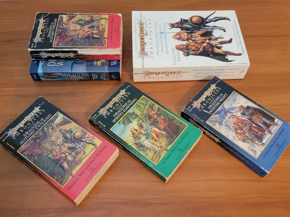 Dragonlance Chronicles Trilogy - paperback books from the 80's, plus the collector's edition, a huge three-book paperback with a white cover. Includes multiple copies of Dragons of Autumn Twilight, Dragons of Spring Dawning, and Dragons of Winter Night. This photo mostly shows the spines, several of which are very good, but one of which (a copy) is tattered.