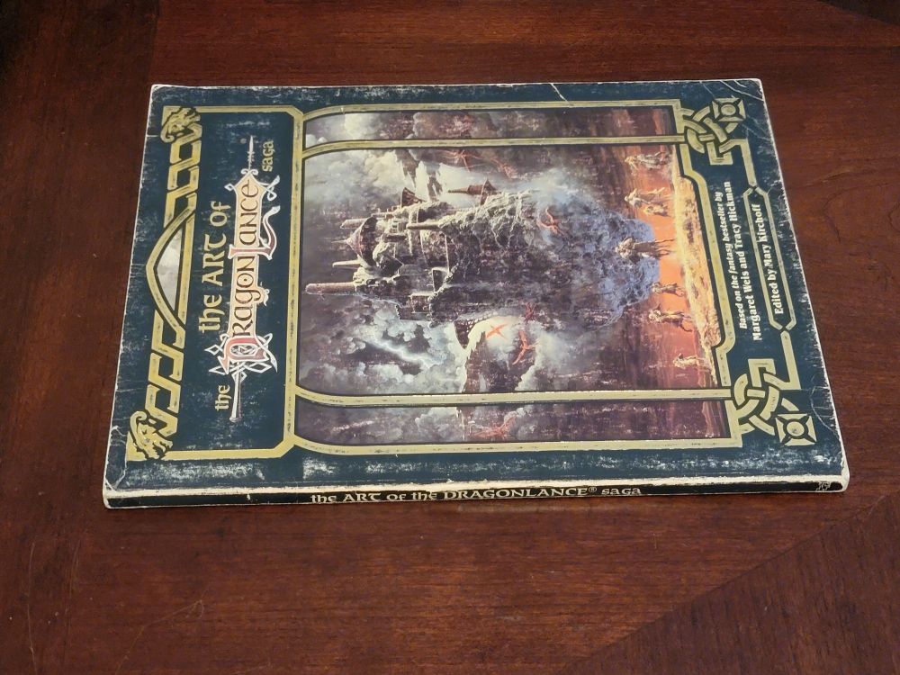 A side view of the Art of the Dragonlance art book, so you can see the spine of this softcover book. The edges are beat up and show white paper through the black background, though there are no splits in the cardboard cover or any tattering or peeling or cracking on the spine. The book is all black and gold with that fantasy painting of a flying castle on the front.