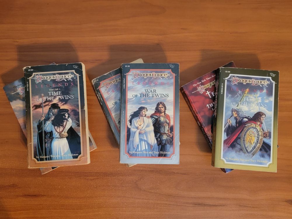 6 Dragonlance paperback novels: the Legends trilogy is in a row on a wooden desk. Each of the three books has another copy of that book underneath: two copies of Time of the Twins, two of War of the Twins, and two of Test of the Twins. The duplicate copies of 1 and 3 are the second edition printings, while the rest are 1st editions. The books are all in good or very good condition.