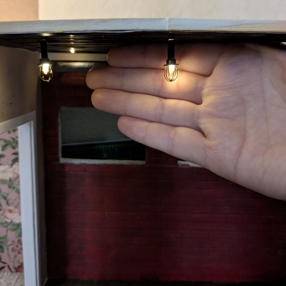 Miniature industrial-style ceiling lighting in a 1:12 scale model house. Two small LED cage lights (sort of look like black, upside-down whisks) emit a warm glow from the black ceiling where they are installed. A person's hand is held behind one of the lighting units to highlight the small scale of the items. In the background, a painted red brick wall with two unfinished window openings. 