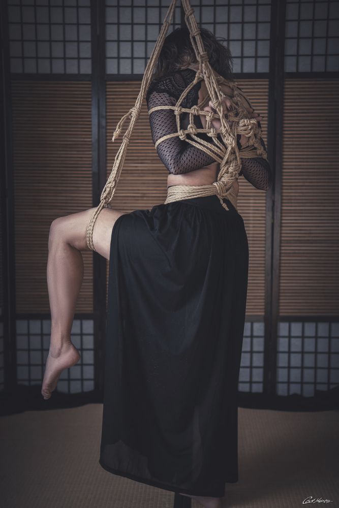 Eine Person ist in einer aufwendigen Shibari-Seilbondage (japanische Kunst des Fesselns) von der Decke hängend zu sehen, fotografiert von hinten. Sie trägt ein schwarzes, transparentes Netzoberteil mit Spitzenmuster und einen langen, fließenden schwarzen Rock. Die naturfarbenen Jute-Seile bilden ein kunstvolles Muster über Rücken, Arme und Oberkörper, während ein Bein leicht angewinkelt ist und das andere gestreckt herabhängt. Der Hintergrund zeigt traditionelle japanische Schiebetüren (Shoji) und Tatami-Matten, was eine ruhige, minimalistische Atmosphäre erzeugt. Das Bild wirkt ästhetisch, elegant und intensiv. Copyright Cat Mason