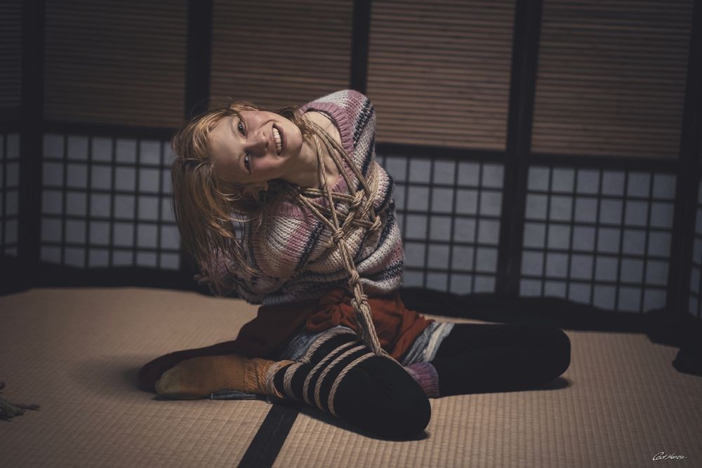 Eine junge Frau mit hellbraunen, leicht zerzausten Haaren sitzt auf einer traditionellen Tatami-Matte in einem japanischen Raum mit Shoji-Schiebewänden im Hintergrund. Sie trägt einen pastellfarbenen, gestreiften Strickpullover, eine rote Hose und schwarze Leggings sowie bunte Socken. Ihr Oberkörper und die Arme sind mit natürlichen Jute-Seilen in einer kunstvollen Shibari-Bindung gefesselt, die über die Brust und um den Hals führt. Die Beine sind leicht angewinkelt und ebenfalls mit Seilen umwickelt. Sie lehnt den Kopf lachend nach hinten, zeigt ein breites, fröhliches Lächeln und wirkt entspannt und vergnügt. Die Szene ist warm und stimmungsvoll beleuchtet. Fotografie von Cat Mason. 

