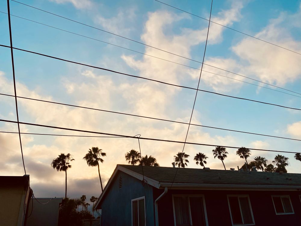 Dark clouds rising up from the west. White clouds in animal shapes becoming golden as the sun sinks behind darkened homes in the foreground. Baby blue behind everything in silhouette. 