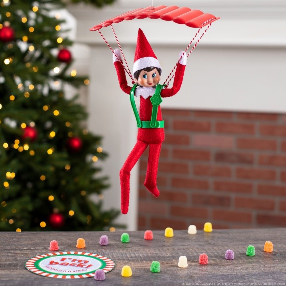 A photograph of the Elf on a Shelf parachuting onto a surface lined with gumdrops (arranged in two parallel lines resembling the landing lights for an airplane). The runway ends in a colorful circle that reads "I'm back!" In the background is a Christmas tree and a brick fireplace.