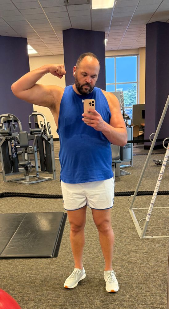 Multiracial man in white shorts and a blue sleeveless shirt flexing his right bicep. 