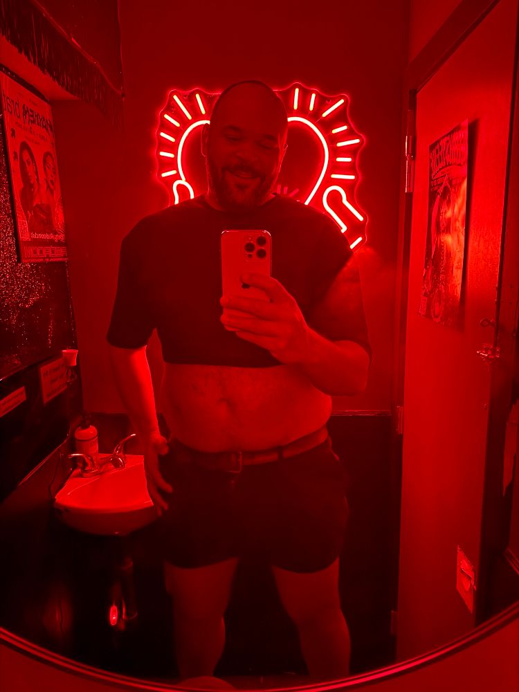 Tall bearish man in a black mesh crop top in a bathroom lit red. 