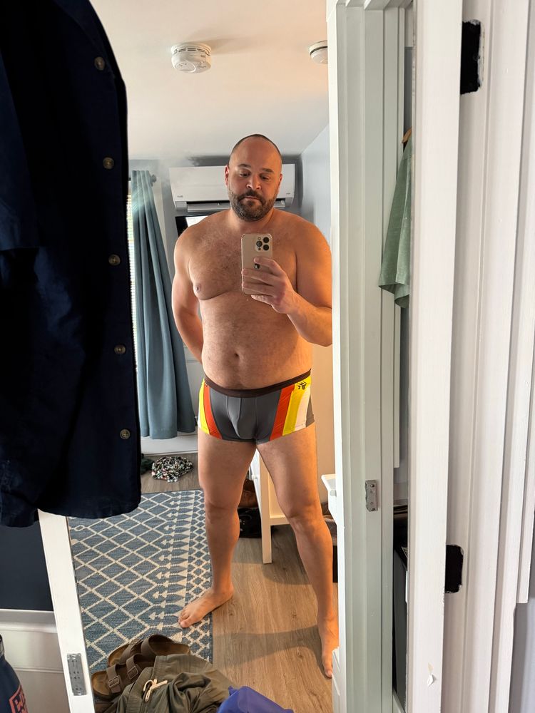 Tall bearish man in a trunk style swimsuit in bear pride colors. 