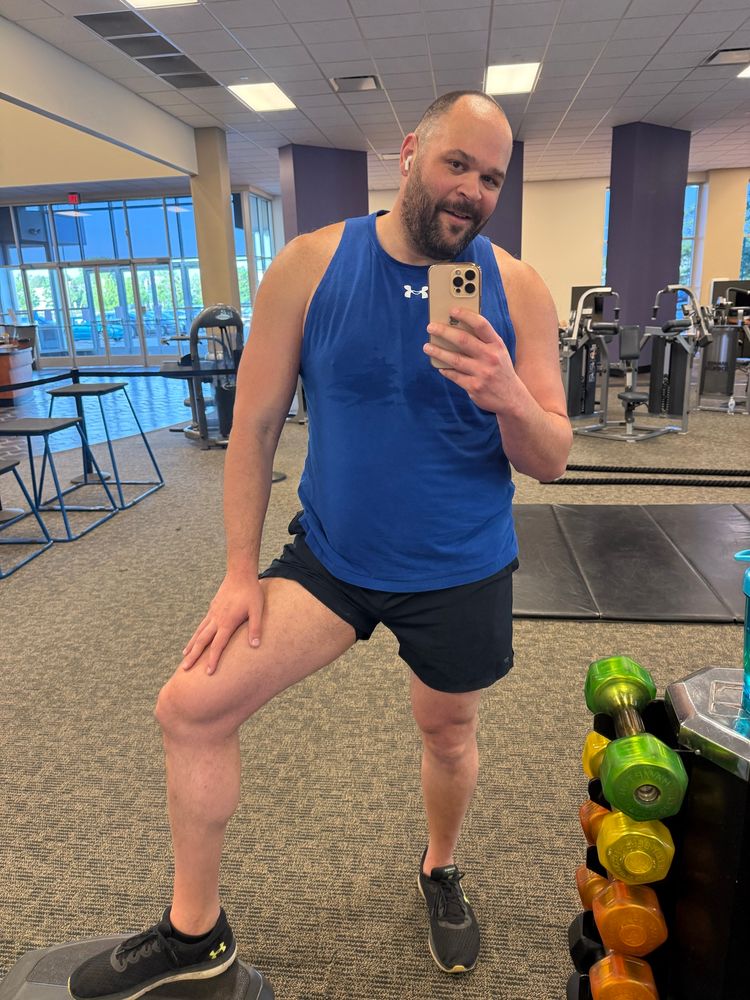 Man in sleeveless blue top and short black shorts in a gym setting with his shorts pull up to show his thigh. 