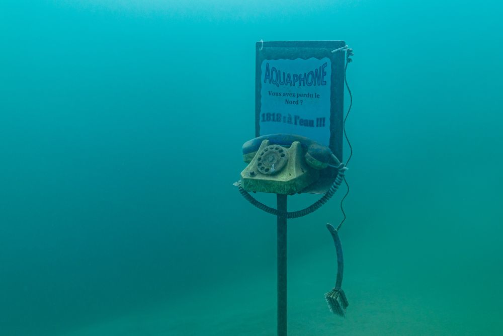 Ein altes Wählscheibentelefon unter Wasser, dahinter ein Schild mit der Aufschrift 