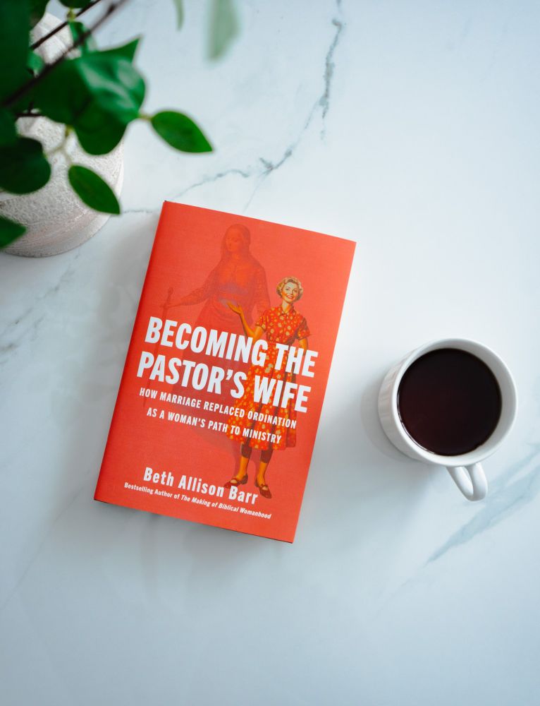 The book is on a white table with a plant and a coffee mug nearby. The book cover is bright pink with the title in bold white letters.