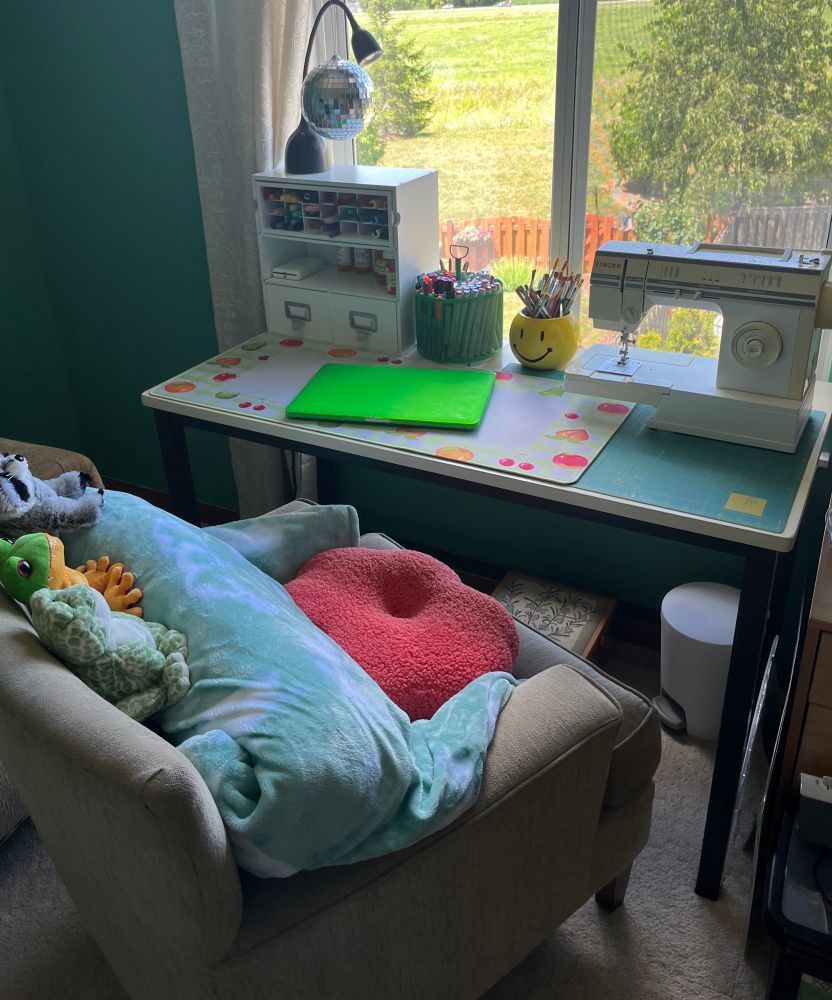 a desk with a comfy lounge chair pushed halfway in, decorated with a pink flower pillow and a tie dye oodie + some webkinz. the desk has some craft supplies, a sewing machine, and multiple craft mats laid down. below the desk is a small white trash can and carpet floors. behind the desk is a window, you can see grass and fence and trees.
