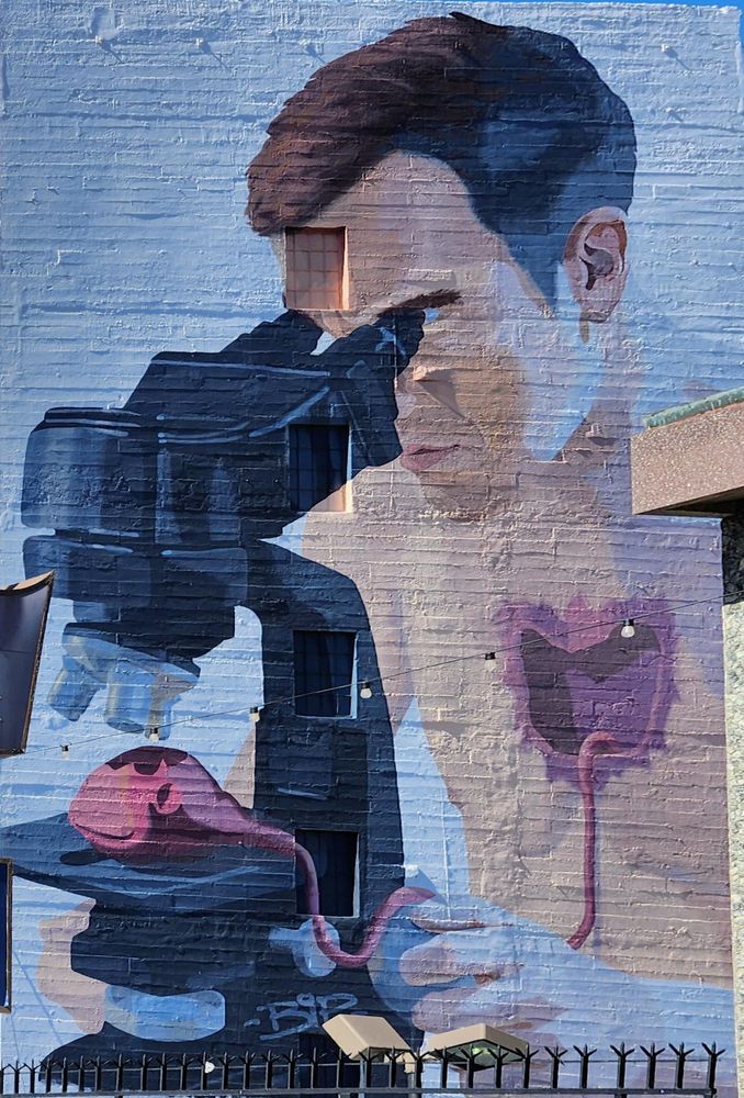 A light-skinned man looks through a microscope at a heart ripped out of his bare chest. A mural painted on the side of a building.