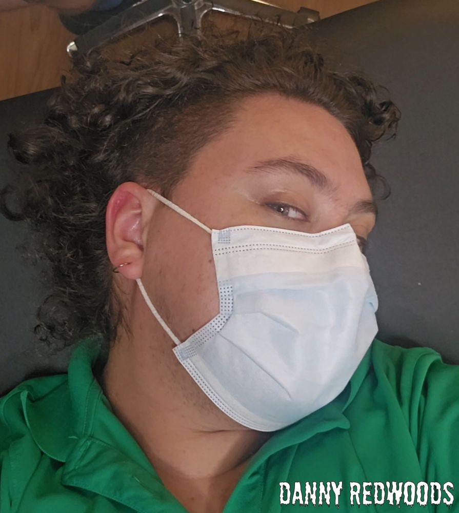 Danny in a green polo shirt and surgical mask, laying on a black table in a piercing shop with his face to the side, looking at the camera