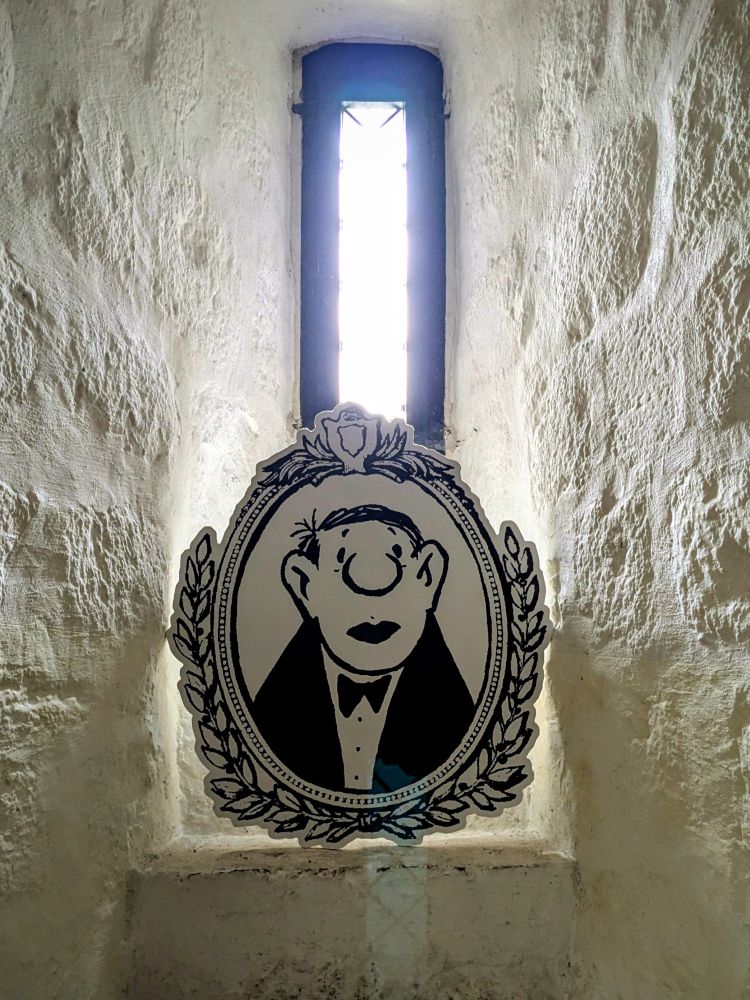 Karikatur von Loriot in einem Lorbeerkranz vor einem schmalen Kirchenfenster.