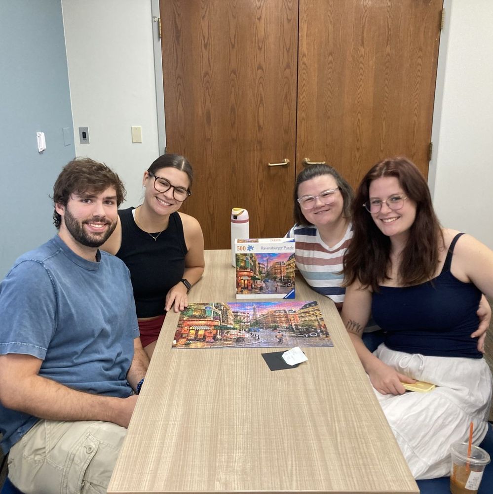 Four people sitting and smiling. On the table is their completed puzzle.
