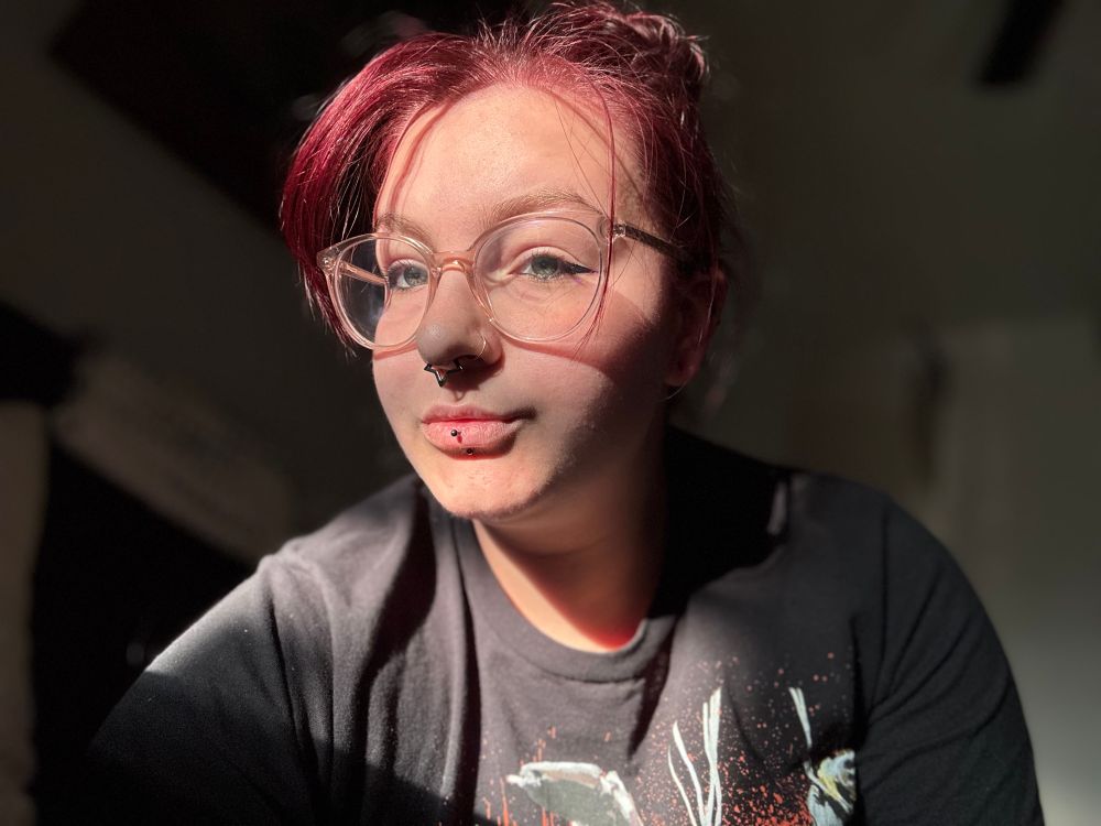 a white femme person with red hair in a bun. they have a nostril piercing, septum piercing, and lip piercing, and clear framed glasses. they are in the sunlight that is highlighting their hair and face