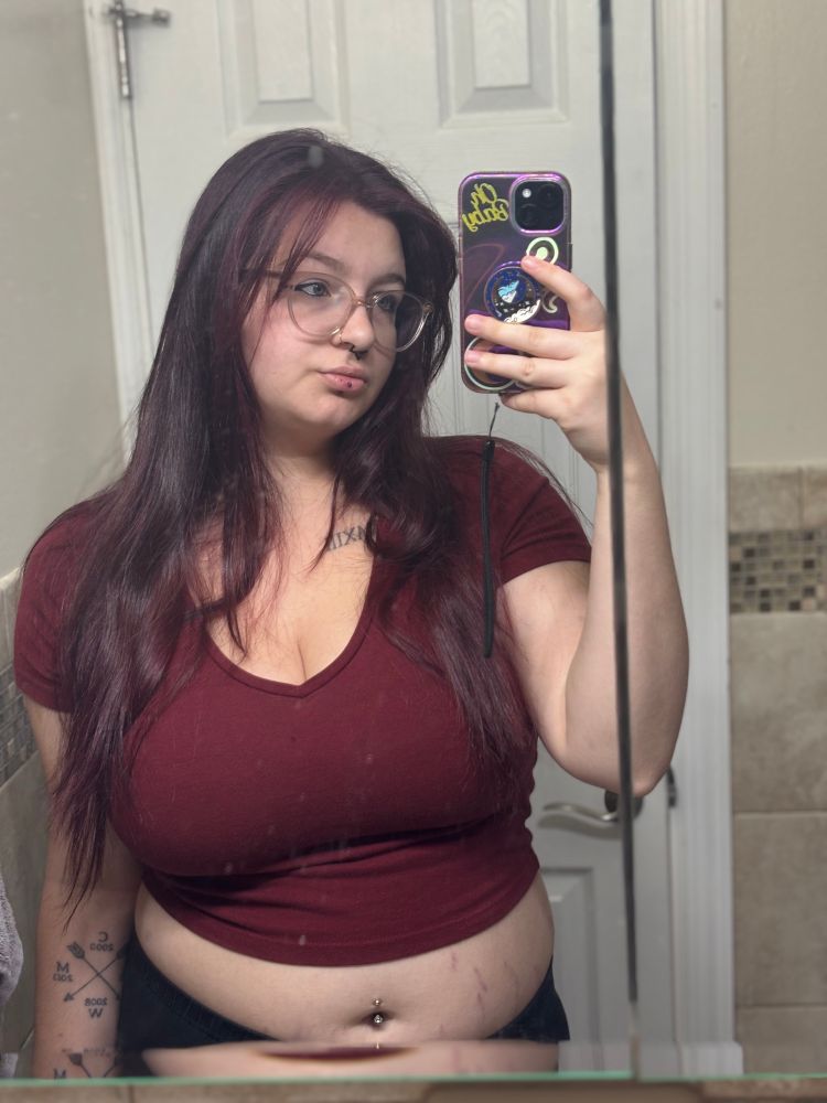 a white femme person with dark red hair, clear framed glasses, nostril piercing, septum piercing, lip piercing, and belly button piercing. they are wearing a  red crop top and black pants. they are taking a selfie in a bathroom mirror