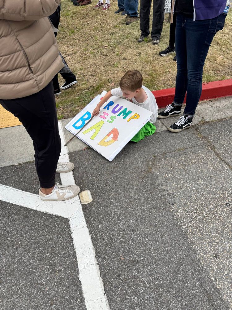 Small boy sitting on the sidewalk at a protest with sign that reads “Trump Is Bad”