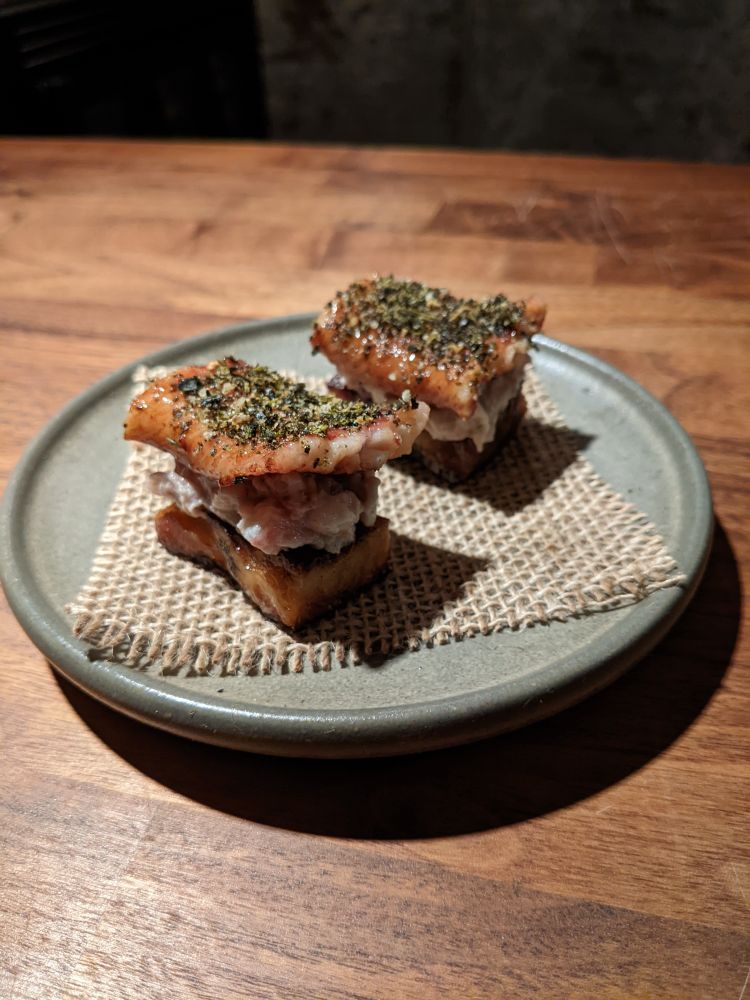 Two small pieces of grilled eel, tolled with green powder, sit on rectangular pieces of a croissant-like cake. They are on a square of burlap, on a round, blue-gray plate, with a wooden counter below.