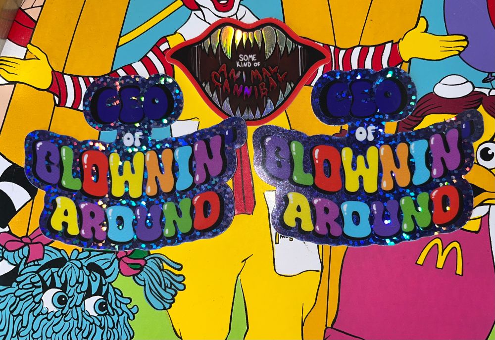 three vibrant stickers from the artist phoxarts against a mcdonald’s placemat. one sticker is a fanged, open mouth with a colorshift finish featuring the lyrics “some kind of animal cannibal” from the horror musical possibly in michigan. the other two say “ceo of clownin’ around” in bright, multicolored bubble letters with a glittery, holographic finish.