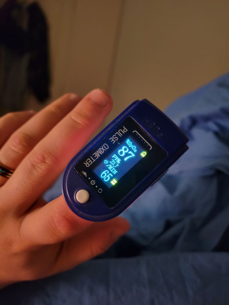 Un saturomètre au bout de mô doigt, affichant une saturation en oxygène de 87 et un rythme cardiaque de 65 battements par minute.