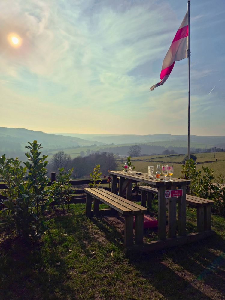 evening sun over a pub garden with the hills of the Peak District in the background. A picnic table with empty beer glasses sits under a waving English flag.