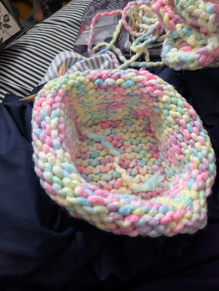 A knitted basket, still on the knitting needles, sitting on a dark blue blanket and black and white striped sheets. In the background, there's a scarf attached to the other end of this basket by the yarn that both are using, it's rolled up, but it is a Leaf pattern scarf.

The basket is made from yarn that is variegated in a rainbow of pastel colors, and a touch of white, giving it a very spring time/Easter/unicorn vomit feeling.