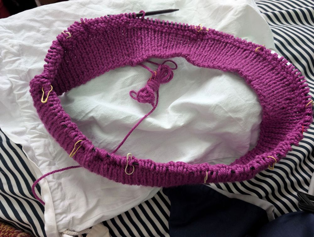The ribbing and beginning of a cowl, on a wooden circular needle, in a darker magenta color, lying on a white cotton bag. 