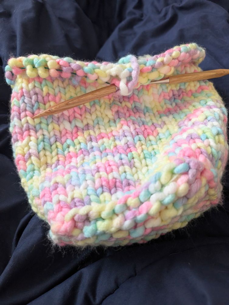 A knitted bag looking object, with a flat base, still on square circular needles, in a bulky sort of pastel rainbow yarn, lying on a dark blue background. 