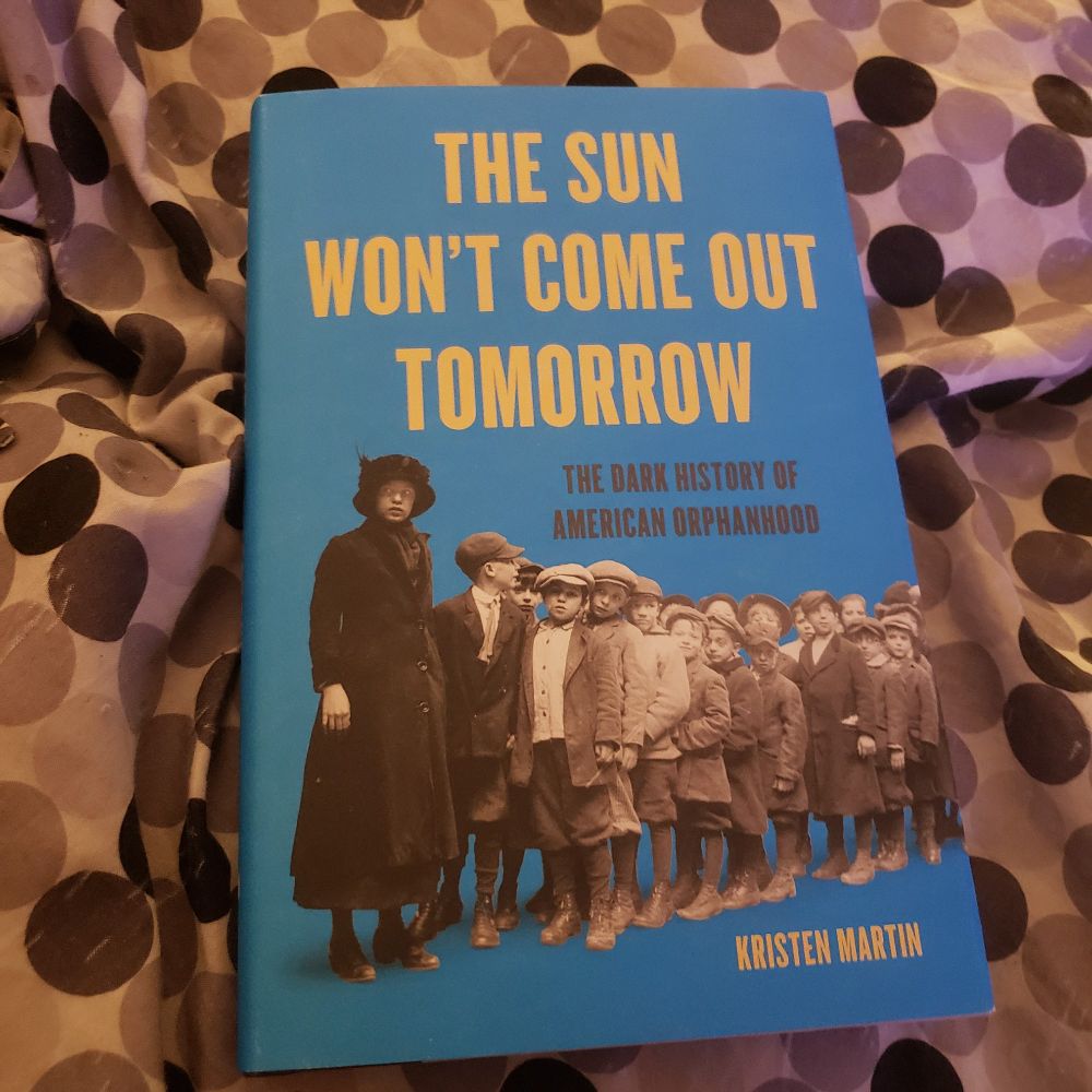 Blue Hardcover Book with photo of orpahns lined up behind a lady, The Sun Won't Come Out Tomorrow: the dark history of American orphanhood by Kristen Martin